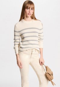 Pull en tricot beige avec des rayures horizontales grises, col rond et poignets côtelés, porté avec un pantalon beige clair et un petit sac à main beige.