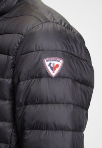 Černá péřová bunda s prošívanými panely a logem Rossignol na rameni. Hladký, lesklý povrch a lehký materiál.