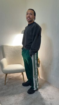 Egy férfi áll egy fehér karosszék mellett bent, fekete pulóvert, zöld Adidas melegítőnadrágot és fekete cipőt visel, kezét a zsebében tartja.