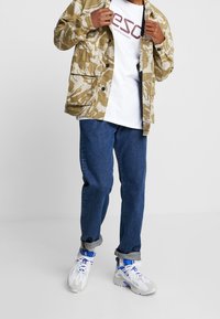 Camo-patterned beige jacket, white t-shirt with red logo, blue denim jeans, and gray/white sneakers with blue accents. Casual outfit.