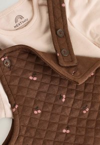 Brązowe pikowane ogrodniczki z różowymi wzorami wiśni, z regulowanymi brązowymi guzikami i jasnoróżową koszulką pod spodem.