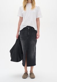 Black denim skirt with front slit, worn with a white t-shirt and slip-on shoes; model holds a black tote bag.