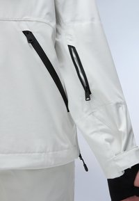 White jacket with black zippers, featuring a textured surface, long sleeves, and adjustable cuffs. Side zipper pockets are visible.