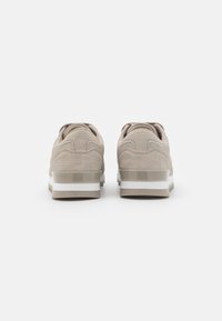 Beige sneakers with a textured upper, padded collar, and white sole. The back features a smooth accent and a patterned material finish.