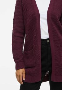 Bordeauxfarbene Strickjacke mit strukturiertem Muster, offenem Vorderteil und zwei Taschen. Gerippte Bündchen und Saum; getragen über einem weißen Hemd und schwarzen Hosen.