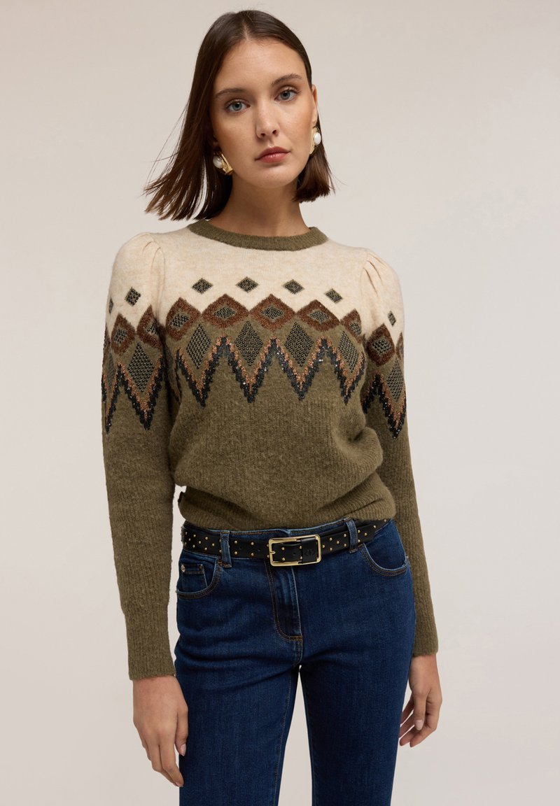 Pulover tricotat de culoare verde măsliniu cu o yoke crem, cu un model ce prezintă forme geometrice. Asortat cu blugi de culoare albastru închis și o curea neagră cu cuie.