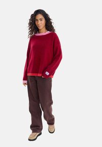 Czerwony sweter z dzianiny z różowym kołnierzem, o luźnym kroju, z prążkowanymi mankietami i dołem. Połączony z brązowymi spodniami o szerokich nogawkach oraz beżowymi butami na sznurowadła.