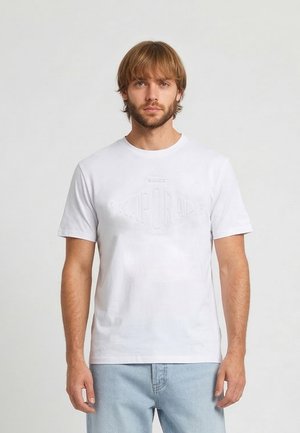 Homme blond avec barbe portant un t-shirt Kaporal blanc uni à manches courtes et un jean bleu clair, debout devant un fond uni gris clair.