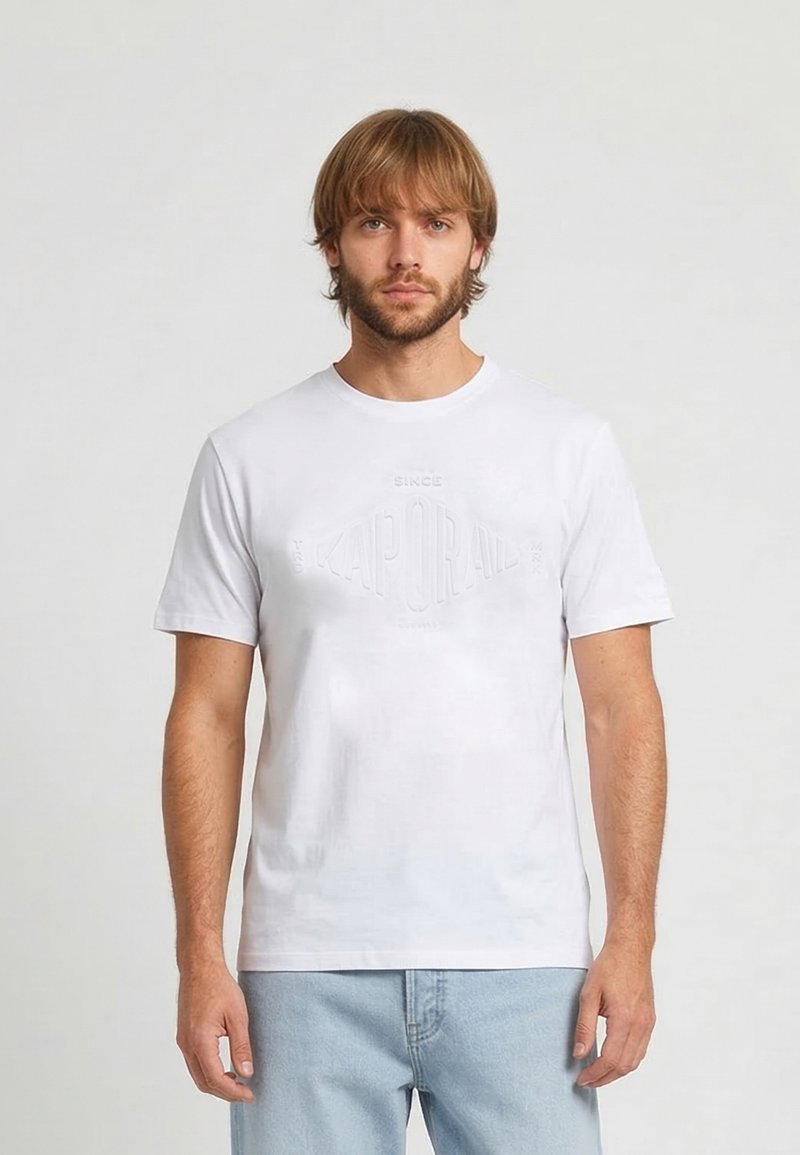 Homme blond avec barbe portant un t-shirt Kaporal blanc uni à manches courtes et un jean bleu clair, debout devant un fond uni gris clair.