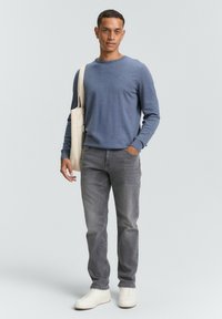 Man in een blauwe trui, grijze spijkerbroek en witte sneakers, met een crèmekleurige draagtas over één schouder, staand tegen een effen achtergrond.