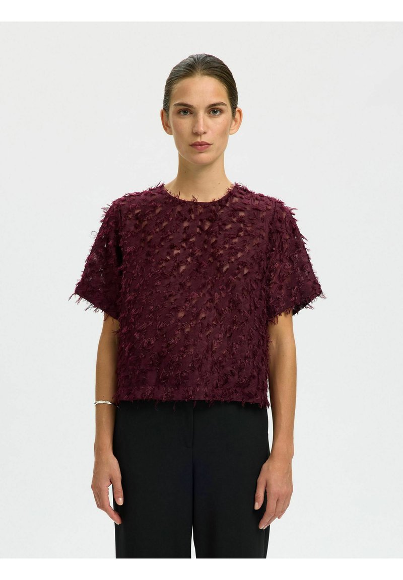 Bordeaux kortemouwen top met textuur en rafelige details; losse, boxy pasvorm; ronde halslijn; gecombineerd met zwarte broeken.
