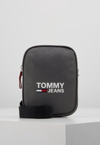 Borsa a tracolla compatta grigia a forma rettangolare con tracolla nera, zip e logo "TOMMY JEANS" con bandiera rossa, bianca e blu sul davanti.