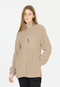 Beige fleecejakke med fuld lynlås, opretstående krave, lynlåslomme på brystet, sidelommer og et logo-patch på venstre side. Glat tekstur.
