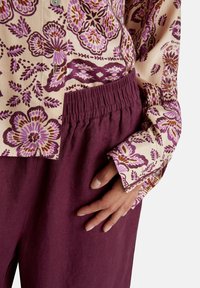 Hand auf burgunderroten, elastischen Bundhosen ruhend, trägt ein beiges Shirt mit lila Blüten- und geometrischen Mustern, ein Knopf ist in der Nähe des Saums sichtbar.