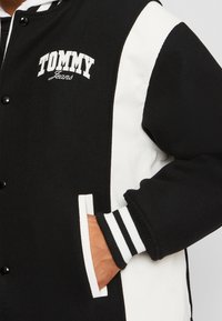 Černobílá varsity bunda s texturovanou tkaninou, s cvoky, bočními kapsami a pruhovanými ribbovými manžetami. Logo na hrudi je viditelné.