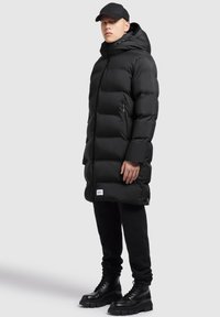 Sort puffjakke med hætte, med lynlås og sidelommer, båret over sorte bukser og sorte snørebåndsstøvler.