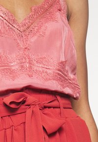 Un débardeur en satin rose avec des finitions en dentelle, présentant un décolleté en V et des bords effilochés. Associé à une jupe rouge plissée taille haute avec un nœud.