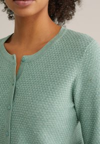 Svetlo zelena kardigan z gumbi, ki ima teksturiran vzorec vafljev, okrogel izrez in gumbe v enaki barvi. Mehki material z nežnim leskom.