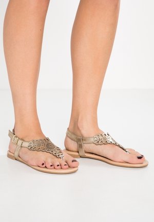 Fødder iført beige, flade sandaler med dekorative udskæringsmønstre og ankleremme på en ensfarvet hvid gulv.