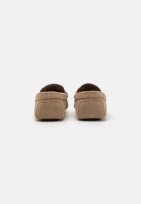 Mocasines de ante en tono marrón claro con un diseño de perfil bajo, parte trasera redondeada y detalles de costura. La superficie texturada y el material suave aumentan la comodidad.