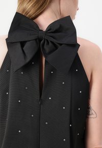 Črna halter majica z velikim vozićem na hrbtu, z teksturirano tkanino in zakovicami na sprednji strani za dodatne podrobnosti.