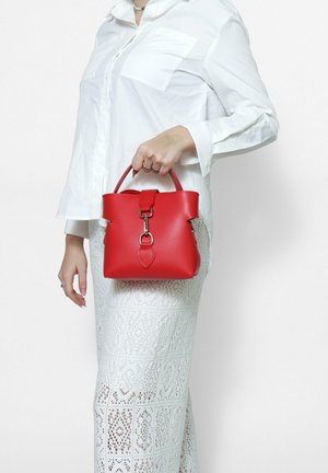 Sac à main en cuir rouge avec une forme structurée, des poignées courtes, du matériel en argent et un accent décoratif en forme de feuille. Détails de surface texturée visibles.