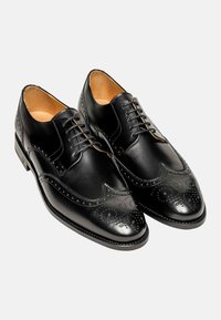 Chaussures noires en cuir avec des détails de brogue, bout pointu et système de laçage. Des motifs texturés accentuent la partie supérieure et les zones des orteils.