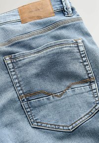 Gai zili džinsi ar brūnu ādas nieri jostasvietā, ar aizmugures kabatu, kas izšūta kontrastējošā dzeltenā krāsā.