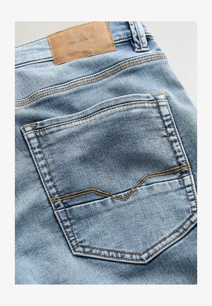 Světle modré džíny z denimu s hnědou koženou nášivkou na pásku, které mají zadní kapsu orámovanou kontrastním žlutým šitím.