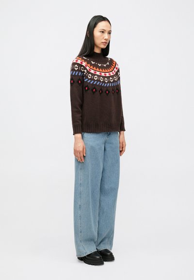 Naine seisab, seljas pruunimustriline kampsun, helesinised laiade säärega teksapüksid ja mustad paksemad kingad, taust on hele ja lihtne.