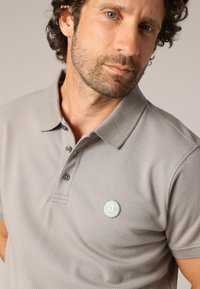 Grijze polo met een gemêleerd patroon, drie knopen aan de voorkant, korte mouwen en een ronde logo-patch op de borst.