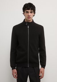 Homme portant un pull zippé noir avec un col montant et deux boutons, se tenant devant un fond blanc uni.