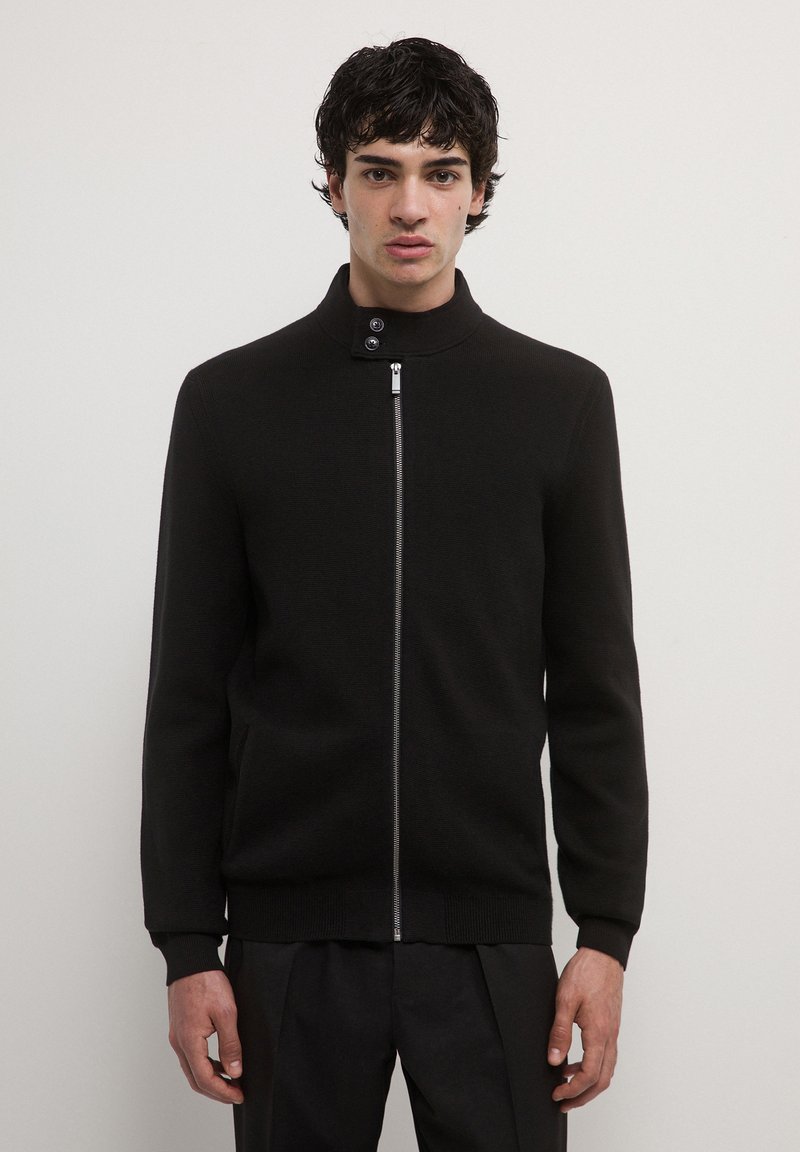 Homme portant un pull zippé noir avec un col montant et deux boutons, se tenant devant un fond blanc uni.