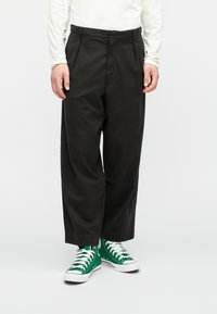 Fekete, könnyű anyagból készült, szabott nadrág, egyenes lábformával és diszkrét ráncokkal, zöld magasszárú sportcipőkkel párosítva.