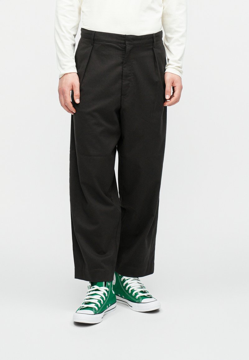 Fekete, könnyű anyagból készült, szabott nadrág, egyenes lábformával és diszkrét ráncokkal, zöld magasszárú sportcipőkkel párosítva.