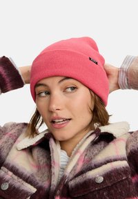 Roze gebreide beanie met een omgeslagen rand en een klein zwart "ROXY" label. Draagt een kleurrijke, geruite fleece jas met drukknopen.