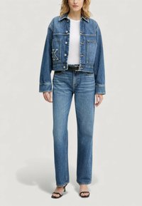 Kurtka jeansowa z logo K na kieszeni, krótki fason, jasnoniebieski kolor, zestawiona ze spodniami jeansowymi o prostych nogawkach, czarnym paskiem i czarnymi sandałami na obcasie.