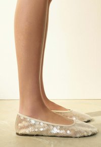 Bēša spīdīgu fliteru baleta čības ar apaļu purngalu, mīkstu tekstūru un nelielu spīdumu, aprīkotas ar plānu apmali ap atvēruma malu.