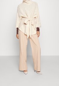 Pull en maille crème avec un col haut et une ceinture à la taille, associé à un pantalon large beige et des chaussures à bout pointu de couleur claire.