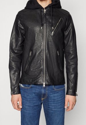 Veste en cuir noire avec fermeture éclair diagonale sur la poitrine, poches avant zippées et capuche noire attachée, portée avec un jean bleu.