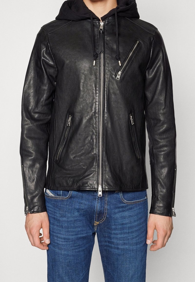 Veste en cuir noire avec fermeture éclair diagonale sur la poitrine, poches avant zippées et capuche noire attachée, portée avec un jean bleu.