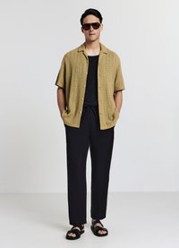 Kurzärmeliges, senffarbenes Strickshirt mit strukturiertem Muster, kombiniert mit schwarzen, schmal zulaufenden Hosen und schwarzen Sandalen. Sonnenbrille verleiht einen modernen Akzent.