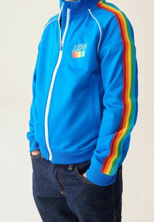 Niebieska kurtka zapinana na zamek z wysokim kołnierzem, białymi akcentami i tęczowymi paskami na rękawach. Noszona z ciemnoniebieskimi dżinsami.