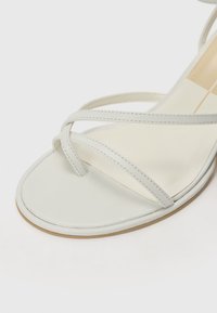 Valkoinen avonainen sandaali ohuilla ristikkäisillä remmeillä ja beige pehmustettu pohja, jossa on ruudullinen kuvio.