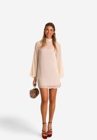 Robe en mousseline de couleur crème, avec de longues manches éventail et un col montant, associée à une pochette beige et des sandales à plateforme rouges.