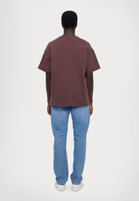 Brązowy bawełniany t-shirt z podwiniętymi rękawami, w zestawie z jasnoniebieskimi jeansami i białymi trampkami, widziany od tyłu.