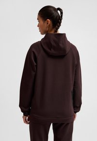 Kobieta odwrócona tyłem, ubrana w ciemnobrązową bluzę z kapturem i dopasowane spodnie, z splecionymi włosami związanymi do tyłu oraz małymi kolczykami w kształcie kółek.