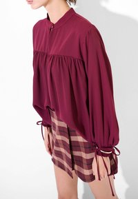 Blusa bordeaux a maniche lunghe con colletto alto e corpetto arricciato, abbinata a una minigonna a quadri nei toni del marrone e del beige.