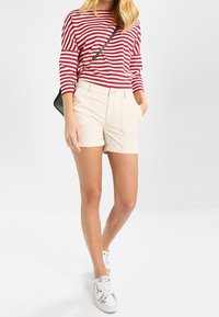 Camisa de rayas rojas y blancas de manga larga combinada con pantalones cortos beige. El modelo lleva zapatillas blancas con un patrón y lleva un bolso negro pequeño.