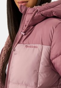 Różowa puchowa kurtka Reebok z pikowanym wzorem, miękkim wnętrzem z polaru oraz regulowanym kapturem ze ściągaczem i zamkiem błyskawicznym.